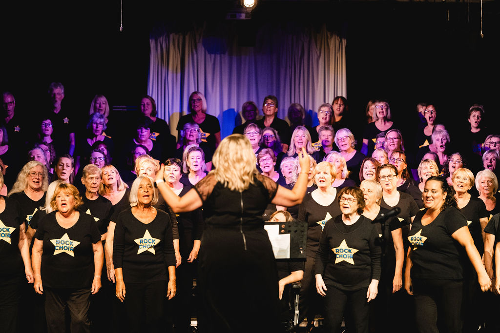 Rows of women dressed in black t-shirts with stars on sing in the Rock choir. They are being led by a woman with their back to the photographer. The choir are performing in a dark walled auditorium, the singers are well-lit under stage lights.