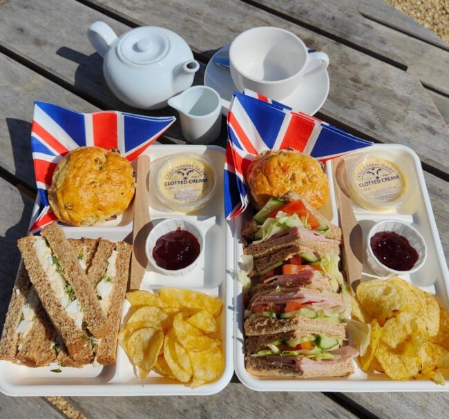 Media (6) Two rectangular plates sit on a wooden table in the Nothe Fort Parade Ground. On the plates, are rows of sandwiches, crisps, and two scones topped with cream and jam. There is a teapot, teacup, and a milk pot also on the table.