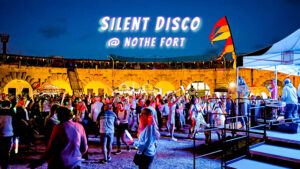 A photo of a previous silent disco at dusk in the parade ground of Nothe Fort. The stone walls are lit up with orange lights and in the centre of the image, a crowd of people are dancing and partying, waving glow sticks and wearing glowing headphones. At the top of the image, across a deep blue night sky, there is a glowy holographic looking font that reads "SILENT DISCO @ NOTHE FORT".