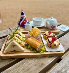 A rectangular plate sits on a wooden table in the Nothe Fort Parade Ground. On the plate, are rows of sandwiches cut into triangles, a sausage roll on a bed of salad, a bowl of crisps, and two scones topped with cream and jam.