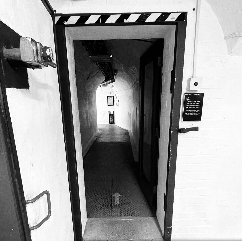 A black and white image of a thick metal door to an emergency nuclear bunker. The corridor beyond the door is dark, but there is a mysterious light at the end of the tunnel.