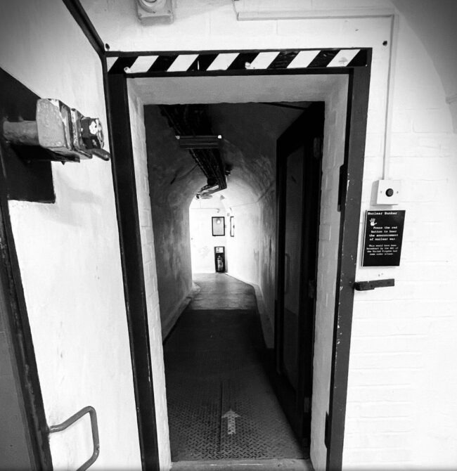 A black and white image of a thick metal door to an emergency nuclear bunker. The corridor beyond the door is dark, but there is a mysterious light at the end of the tunnel. The Text reads COLD WAR BUNKER TOURS