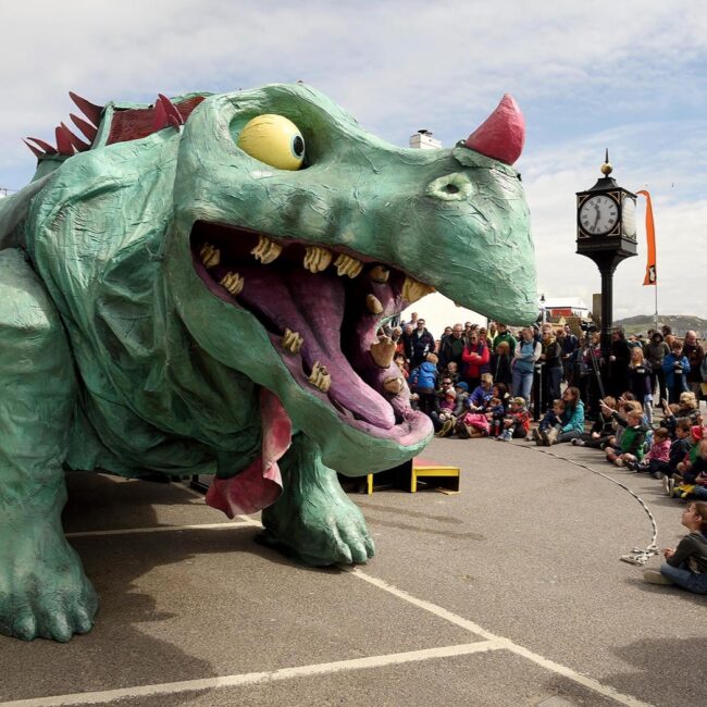 A Large green and red iguanodon dinosaur constructed from recycled materials stands tall in front of a crowd of families. The scene is a street performance.