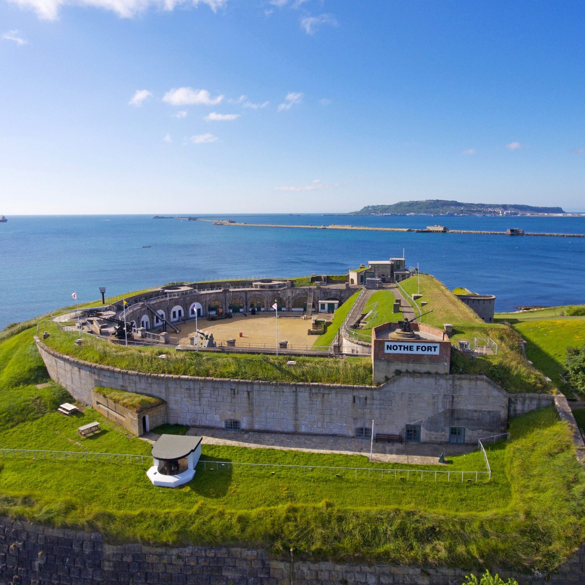 An aerial photo of Nothe Fort, with a clear blue sky above and the sea beyond contrasting with the bright green verges of the D shaped Fort.