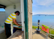 A photo taken from outside the signal lighr room. Half od the image show inside the room, with our Artist in Residence hard at work, working on a painting on a table. Mala is looking at the camera and smiling with sunglasses on. The right hand side of the images shows the view from the Ramparts, capturing the amazing views surrounding Nothe Fort of Portland, the breakwater, and the blue sky and sea beyond.