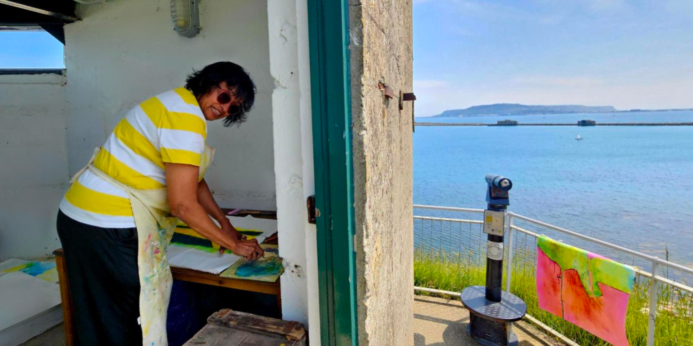 A photo taken from outside the signal lighr room. Half od the image show inside the room, with our Artist in Residence hard at work, working on a painting on a table. Mala is looking at the camera and smiling with sunglasses on. The right hand side of the images shows the view from the Ramparts, capturing the amazing views surrounding Nothe Fort of Portland, the breakwater, and the blue sky and sea beyond.
