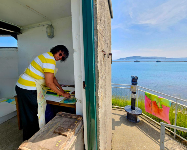 Mala in the Signal light room A photo taken from outside the signal lighr room. Half od the image show inside the room, with our Artist in Residence hard at work, working on a painting on a table. Mala is looking at the camera and smiling with sunglasses on. The right hand side of the images shows the view from the Ramparts, capturing the amazing views surrounding Nothe Fort of Portland, the breakwater, and the blue sky and sea beyond.
