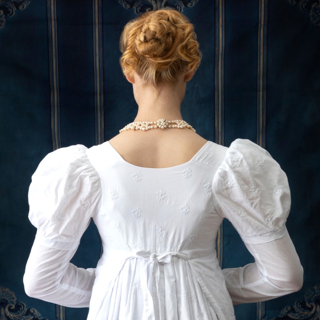 A woman in a white regency era dress stands with her back to the viewer. Behind her is a dark blue decorative wallpaper.