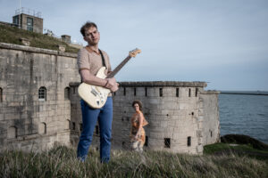 Two performers stand outside the stone walls of the Fort. One is in the foreground holding a white electric guitar, and the other stands further in the background in an orange dress. 