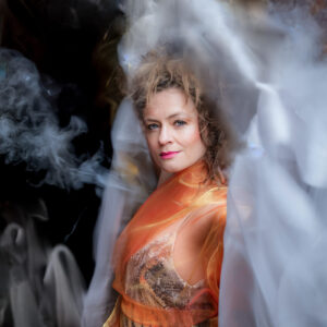 A performer in a shiny orange dress stands in a mysterious smoky mist against a dark black background.