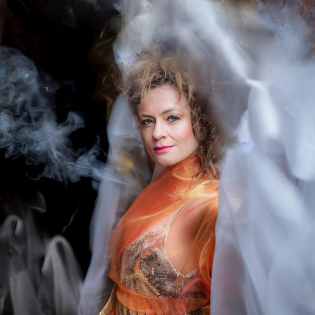 A performer in a shiny orange dress stands in a mysterious smoky mist against a dark black background.