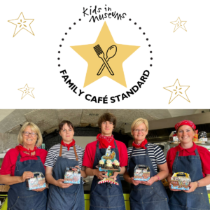 Kids museums cafe One half of the square image includes the kids in museums font logo at the top of a circle of dots, in the middle is a gold 5 pointed star with crossed kitchen utensils in black lined art. The writing at the bottom of the circle reads FAMILY CAFE STANDARD. Beneath that is an image of five of the cafe team holding trays of cakes and example lunch bags.