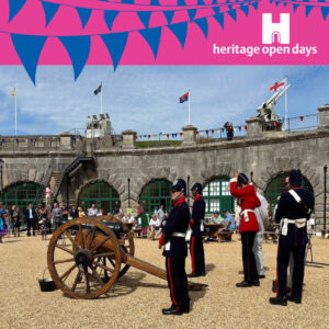 A square image of a photo of the Artillery in Nothe Fort posing with their cannon. Above the blue sky of the image is a bright pink banner with blue bunting across the logo. In the right hand corner of the pink header is the white heritage open days logo.