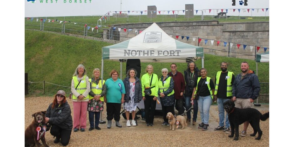 A line up of 12 human volunteers and staff, and three fine dogs represent the Love Dogs West Dorset and Nothe Fort teams. Behind them is a Nothe Fort white and green gazebo, and the walls of the Fort. Above them in blue text reads a thank you message.