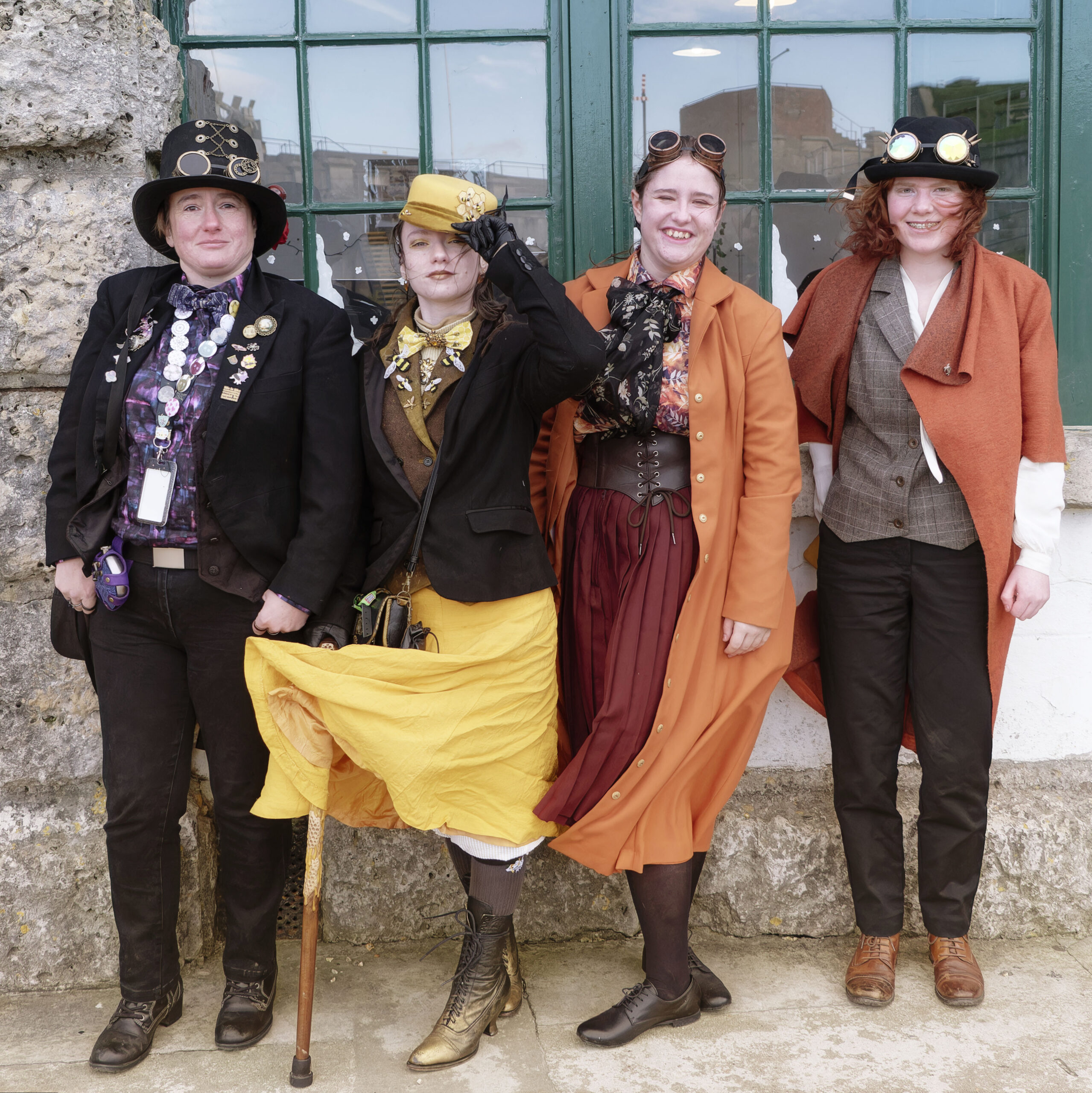 Four well-dressed steampunk individuals, with Victorian inspired attire with an industrial twist: top hats, bedazzled goggles, canes, and corsets.