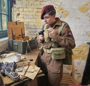 An image of a young man dressed as a WW2 soldier using radio equipment. All around him are authentic looking props. He is standing inside one of the Fort's WW2 education rooms.