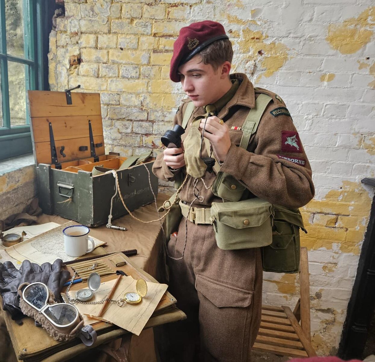An image of a young man dressed as a WW2 soldier using radio equipment. All around him are authentic looking props. He is standing inside one of the Fort's WW2 education rooms.