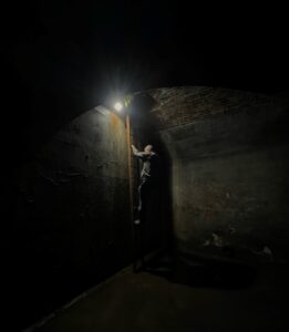 An eerie dark photo of someone descending a ladder with a torch into the water tank. The image edges are surrounded in shadows.
