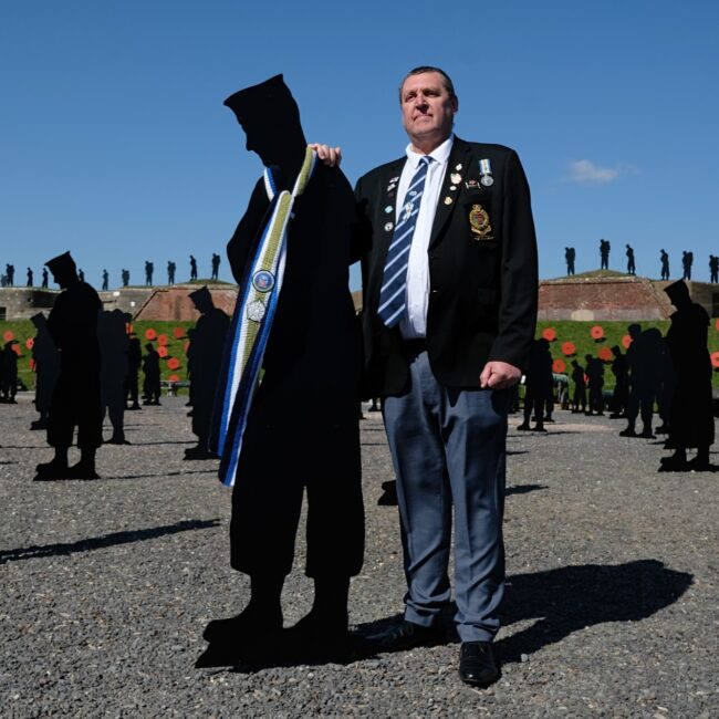 A Falklands veteran stands proudly in a black suit beside one of the metal silhouettes of the 255 forces personnel who were lost in the Falklands conflict. They both stand in the parade ground. In the background are many more black metal silhouettes standing on display.