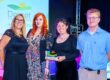 Four Nothe Fort employees stand in a row on stage. The first figure is wearing a smart black dress and has blonde hair. The second is wearing a multicoloured patterned dress and has long red hair. The third staff member is wearing a sparkly black top and long skirt. They are holding the glass award with the rolling hills logo of the Dorset Tourism Awards. The Fourth member of staff is in a light blue shirt and dove grey trousers.