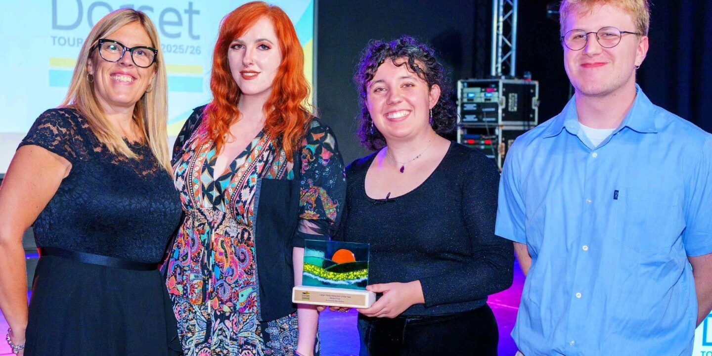 Four Nothe Fort employees stand in a row on stage. The first figure is wearing a smart black dress and has blonde hair. The second is wearing a multicoloured patterned dress and has long red hair. The third staff member is wearing a sparkly black top and long skirt. They are holding the glass award with the rolling hills logo of the Dorset Tourism Awards. The Fourth member of staff is in a light blue shirt and dove grey trousers.