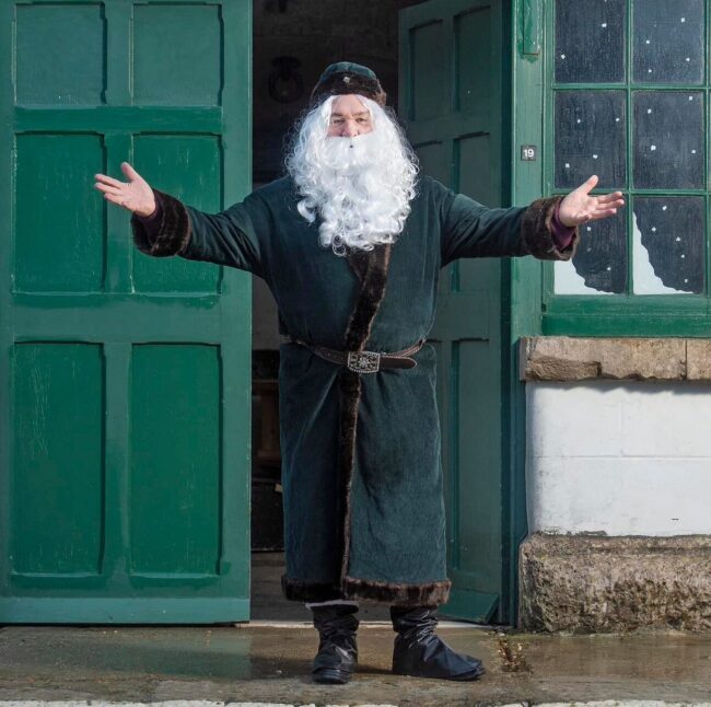 A traditional victorian father christmas wearing a green suit with a dark green hat stands in front of the bottle green doors of the Fort's family Room. His arms are outstretched in welcome. He has a long white beard.