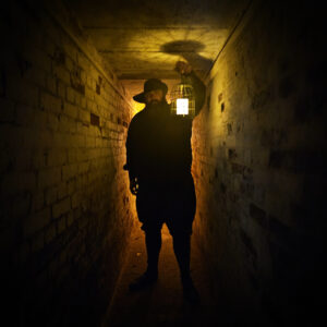 An image of dark figure almost in shadow stands in a spooky looking tunnel. He is carrying an old candle lantern that casts a yellow glow on the bricks above and around him. The figure is backlight by a soft orange light.