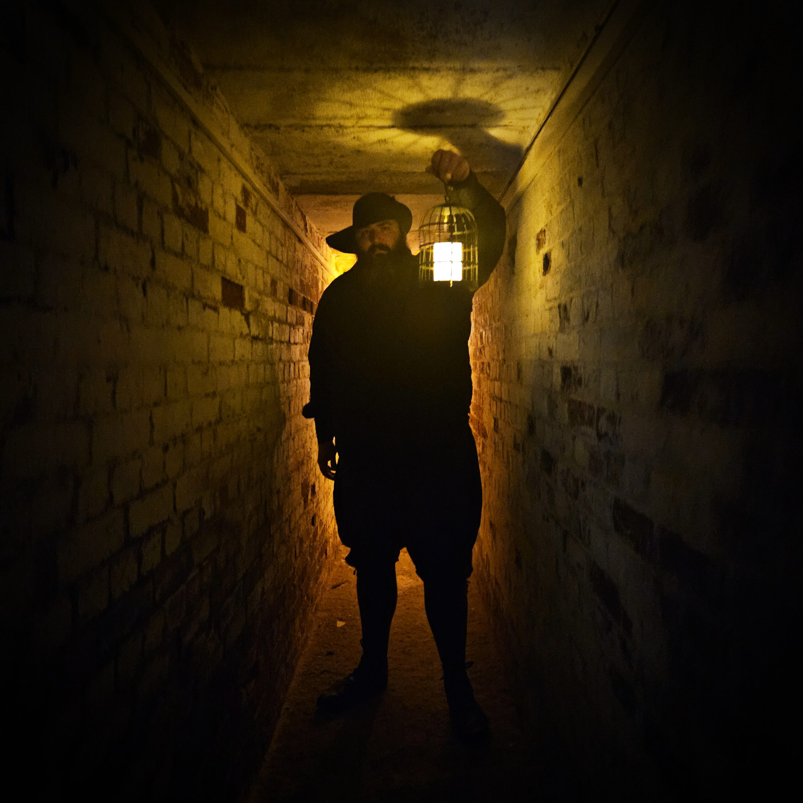 An image of dark figure almost in shadow stands in a spooky looking tunnel. He is carrying an old candle lantern that casts a yellow glow on the bricks above and around him. The figure is backlight by a soft orange light.
