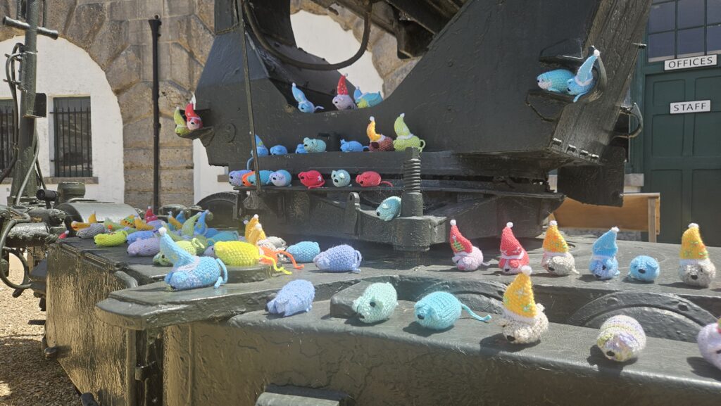 A photo of many multicoloured knitted mice sitting on the large search light in the parade ground. 