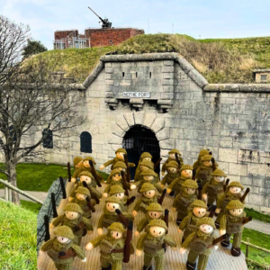 A knitted panel of five rows of WW2 soldiers sits in front of Nothe Fort to demonstrate the exhibition is coming to the Fort. In the background, the grey stone walls of Nothe Fort and the dark archway of the main gate. Above the wall on the ramparts, a heavy artillery gun is visible.