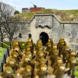 A knitted panel of five rows of WW2 soldiers sits in front of Nothe Fort to demonstrate the exhibition is coming to the Fort. In the background, the grey stone walls of Nothe Fort and the dark archway of the main gate. Above the wall on the ramparts, a heavy artillery gun is visible.