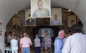 A photo of the Amnesty Exhibition launch party. Shows a large vaulted ceiling room in the Fort with banners of photographed subjects hanging down from the ceiling. A crown of people are gathered within the room.