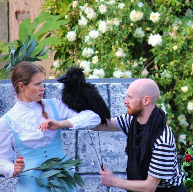 A photo of two actors from the Alice in Wonderland show. One performer is dressed in the blue and white dress of Alice, and is holding a crow puppet on her arm. Another performer is an inhabitant of Wonderland, is puppeteering the crow.