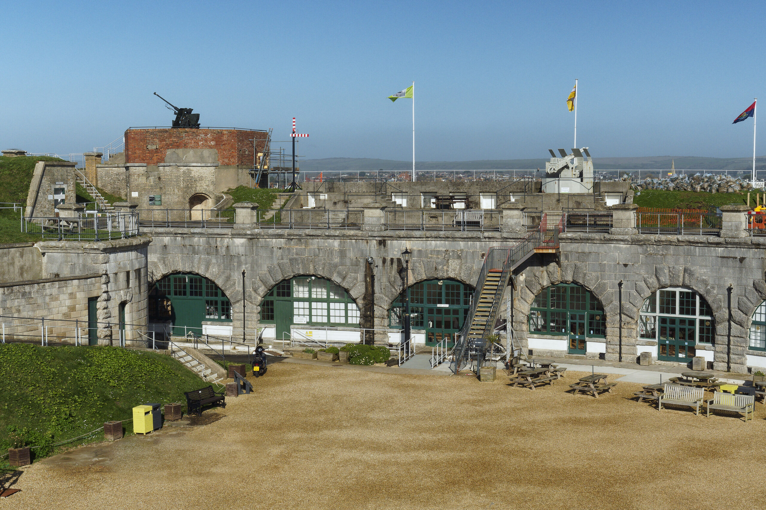 A very similar view of the Fort casemates and ramparts taken in 2024 to contrast with the older images of the Derelict Fort in the 1970s. The parade ground and ramparts are tidy, clear of rubbish, grass is cut and the three flagpoles are adorned in flags blowing in the wind against a blue sky.