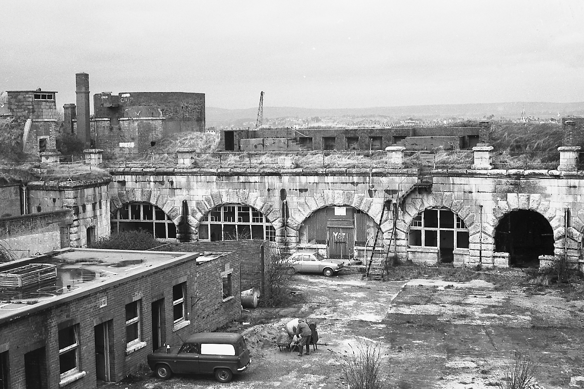 A black and white photo taken in 1970's of the derelict Fort showing shattered glass of the circular windows of the casemates inside the Fort's wall's. There is plenty of debris and overgrowth of weeds around the ramparts and parade ground. Taken by Stuart Morris.