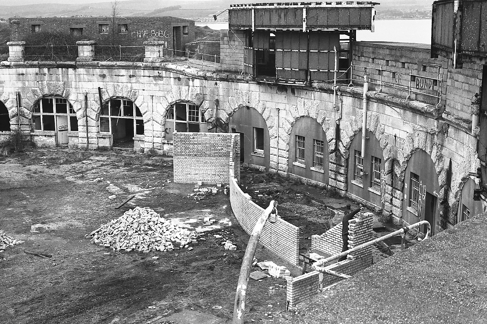 A black and white photo taken in 1970's of the derelict Fort showing shattered glass of the circular windows of the casemates inside the Fort's wall's. There is plenty of debris and overgrowth of weeds around the ramparts and parade ground. Taken by Stuart Morris.