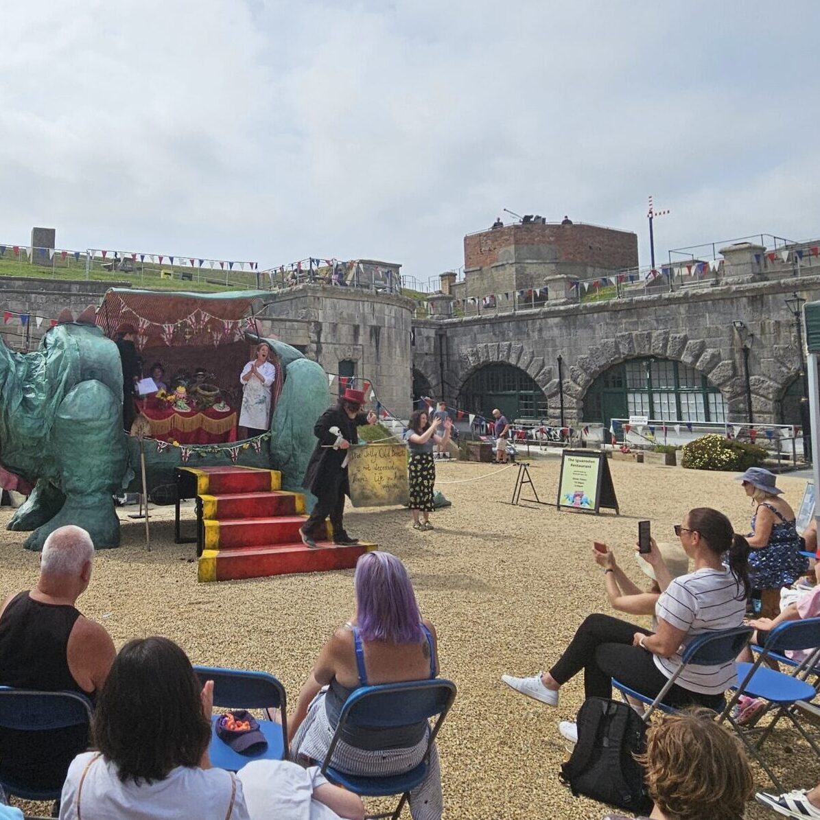 A photo of Fossil Fest weekend and performances of Emerald Ant's Iguanodon Restaurant which involves a large green iguanodon set piece.
