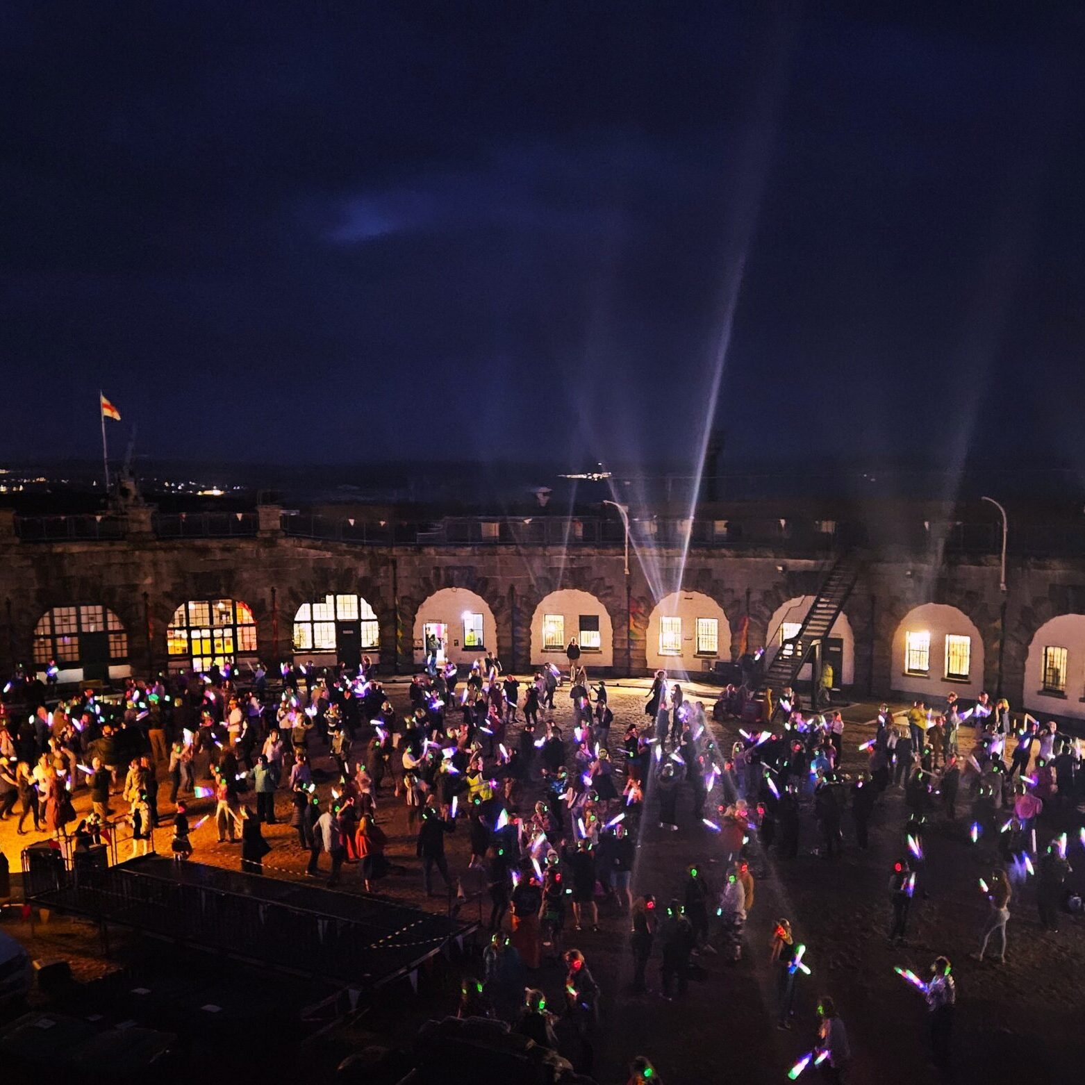 A photo of the 2025 summer silent disco. A large crowd dances in the dark while the white lights of the circular Fort's walls illuminate the parade ground.