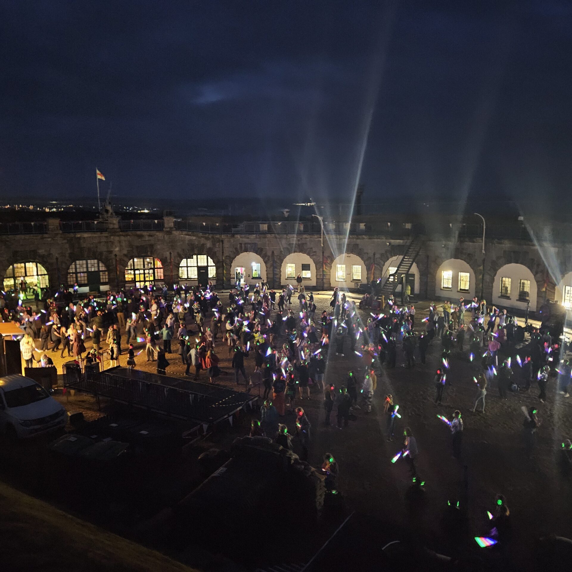 A photo of the 2025 summer silent disco. A large crowd dances in the dark while the white lights of the circular Fort's walls illuminate the parade ground.