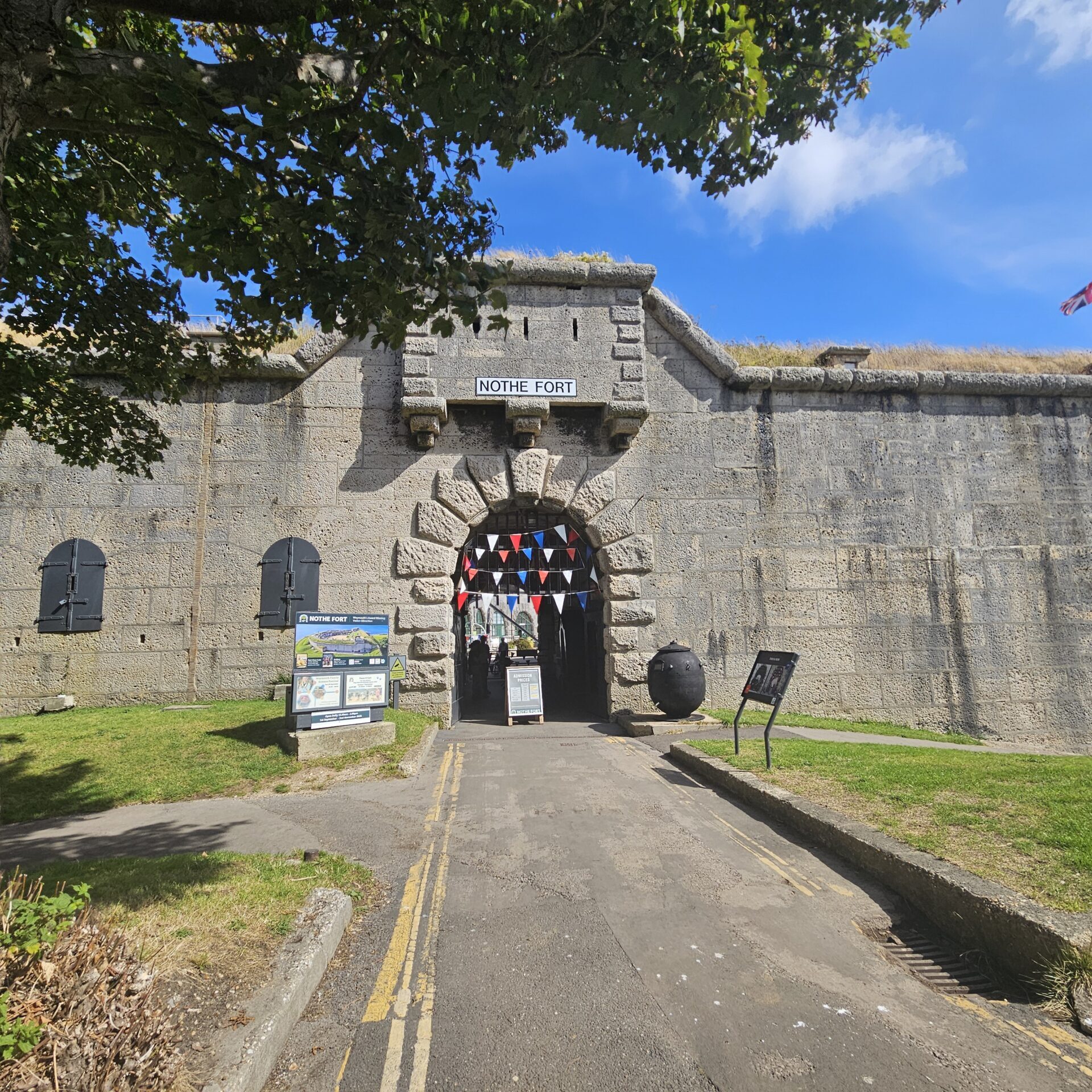 A photo of the entrance tunnel to the Fort, taken outside on a bright sunny day. There is a green verge running either sides of the entrance tunnel arch, with a leafy green tree to left of the tunnel. The portcullis has red white and blue bunting decorating it, and there are two information boards outside the Fort as well as a decorative Sea Mine.