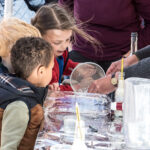 A photo of a previous science fair event with a group of children crowding around a table to see a water-based experiment.