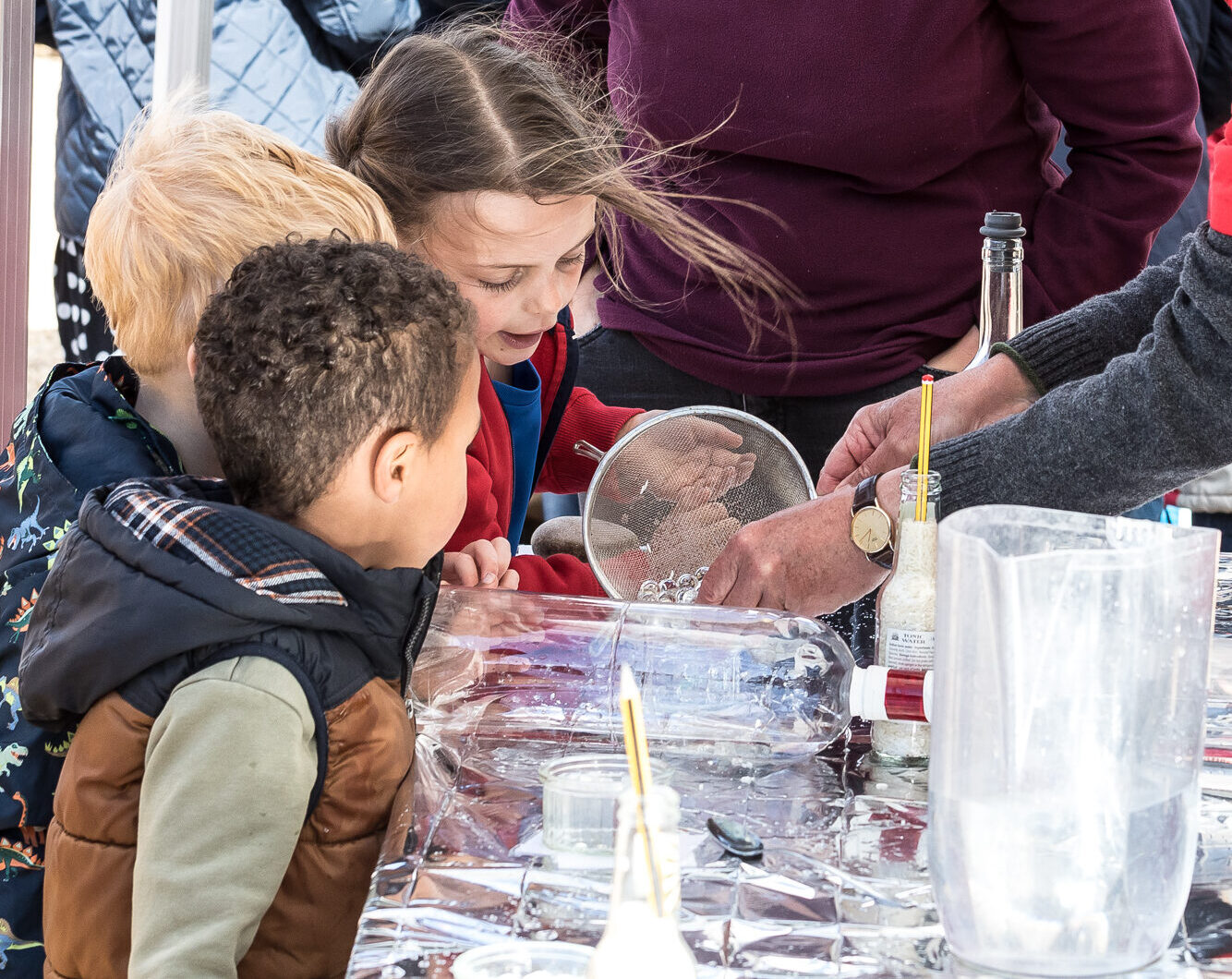 A photo of a previous science fair event with a group of children crowding around a table to see a water-based experiment.