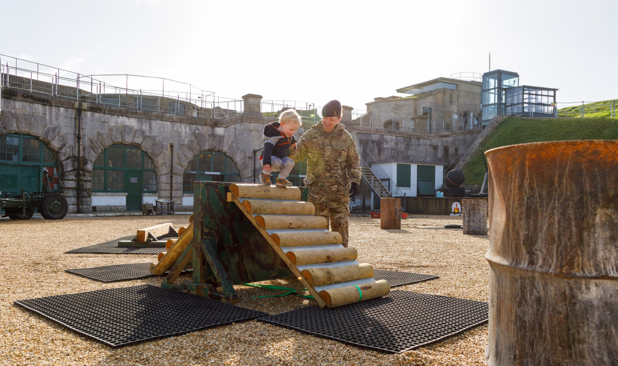 A man in uniform helps a young girl climb the assault course bridge obstacle in the Parade Ground. In the background more fun obstacles can be seen. Photo credit Paul Brewer