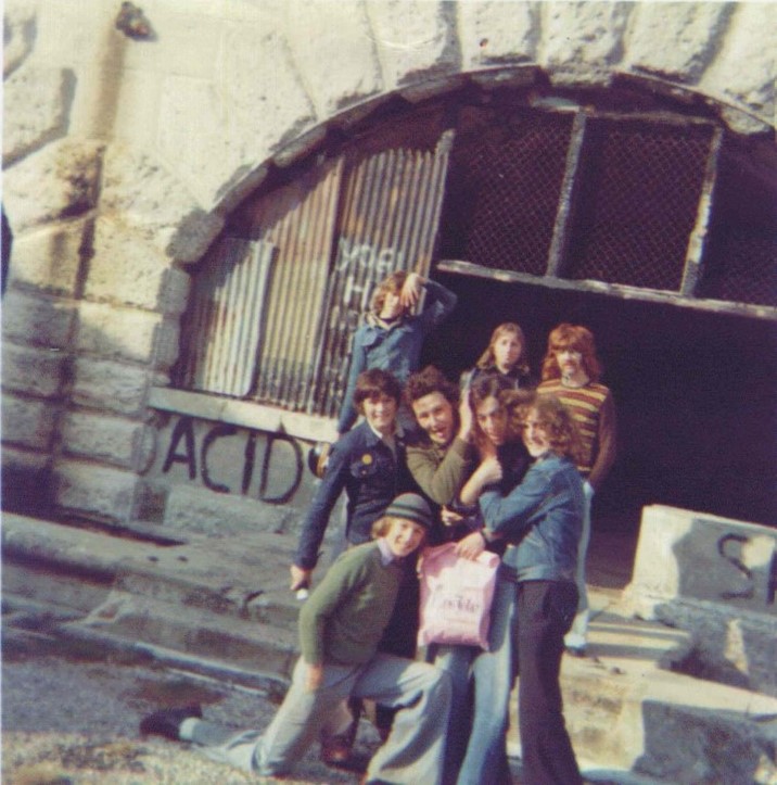 An old photo of a group of young rebels pose together for a photo in front of a graffiti covered arched window in the Fort.