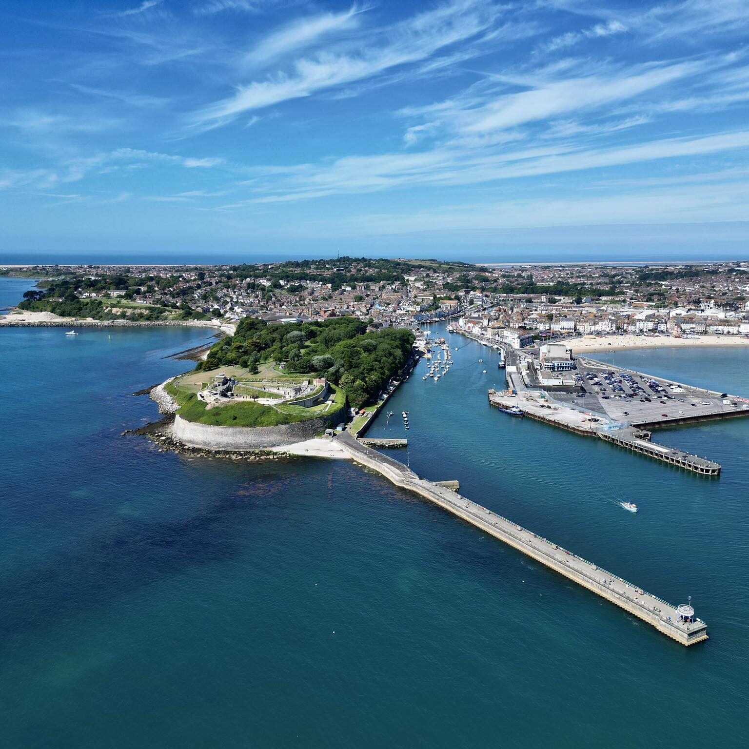 An aerial photo of Nothe Fort, with a clear blue sky above and the sea and Weymouth town beyond contrasting with the bright green verges of the D shaped Fort.