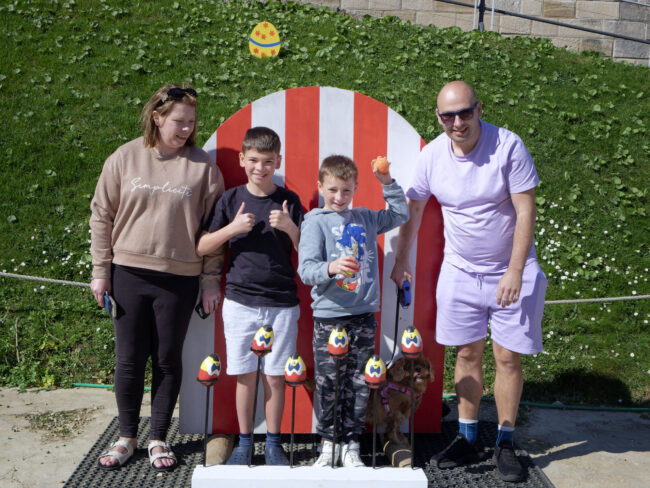 family of four stand in the parade ground in front of an Easter themed coconut shy throwing game.
