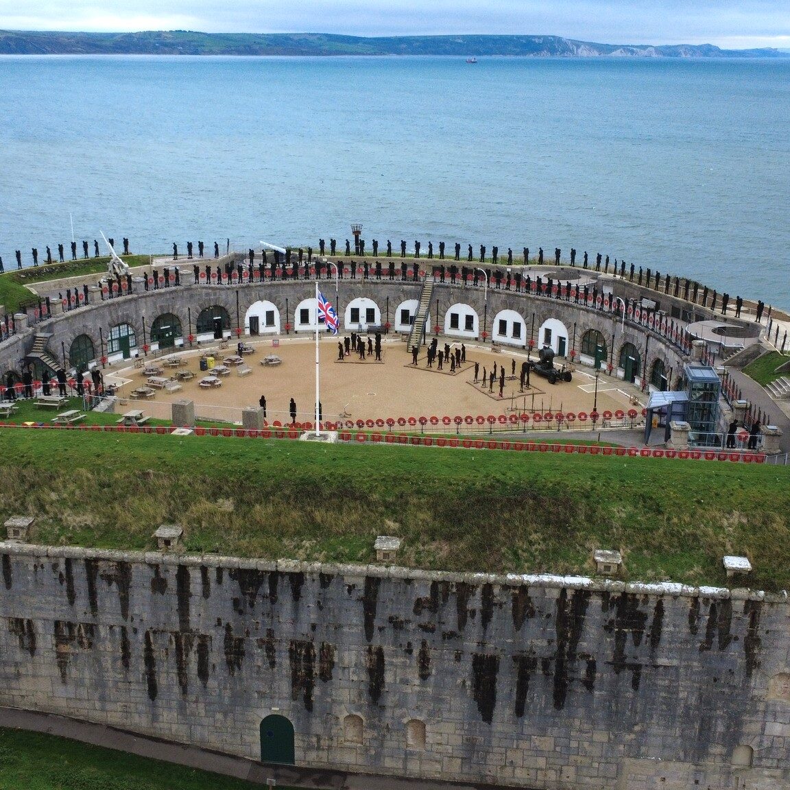 Drone Photo by Eon Matthews 2025, an aerial photo taken from the flat wall side of the D-shaped Fort. The curved walls of the Fort's parade ground can be seen. In the distance is Weymouth Bay. Lining the walls of the Fort are the 200+ silhouette figures that make up the Standing With Giants Falklands Tribute.