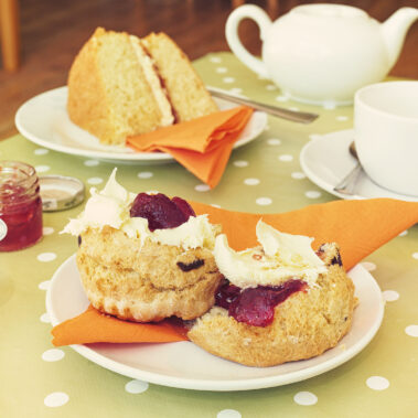 A photo of an afternoon tea spread on a table, with teapot, scones with jam and cream, and a slice of Victoria sponge cake.