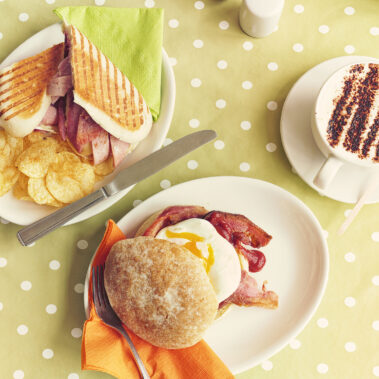 A photo of two plates of delicious hot food options, a bacon and bree panini with crisps, and a bacon and egg bap with a coffee.