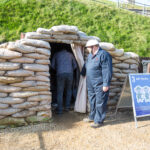 A man in a blue boiler suit is wear a black and white ARP warden tin hat in the style of WW2 era. He is holding a curtain back of the fort's anderson shelter, which is built from stacked sandbags and sheet metal. Behind the shelter is a grassy hill.